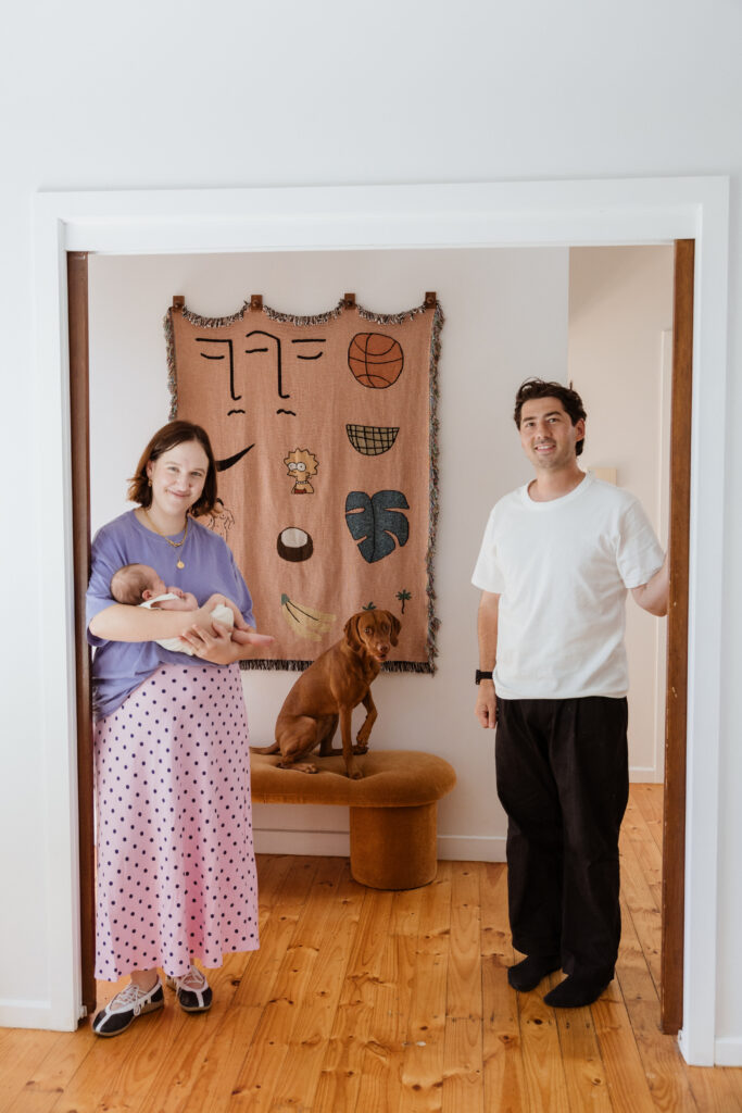 Woman holding a baby stands on the left of a doorframe and a man to the right. In the background a dog sits on a bench.