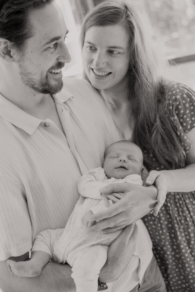 black and white closeup of dad holding newborn while mum stands close and smiles at him