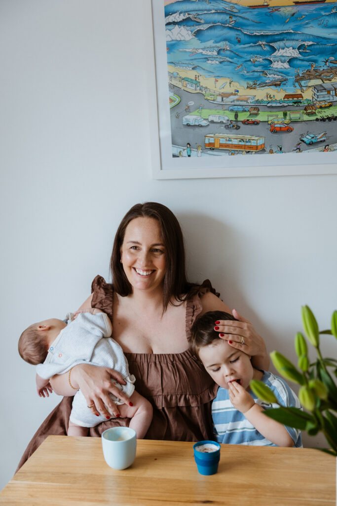 mum holds newborn while sitting at dining table with toddler, stroking his head as he eats biscuits during in-home newborn session