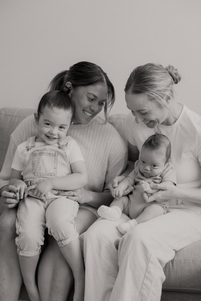 Two mums holding toddler and newborn on their laps, smiling at each other
